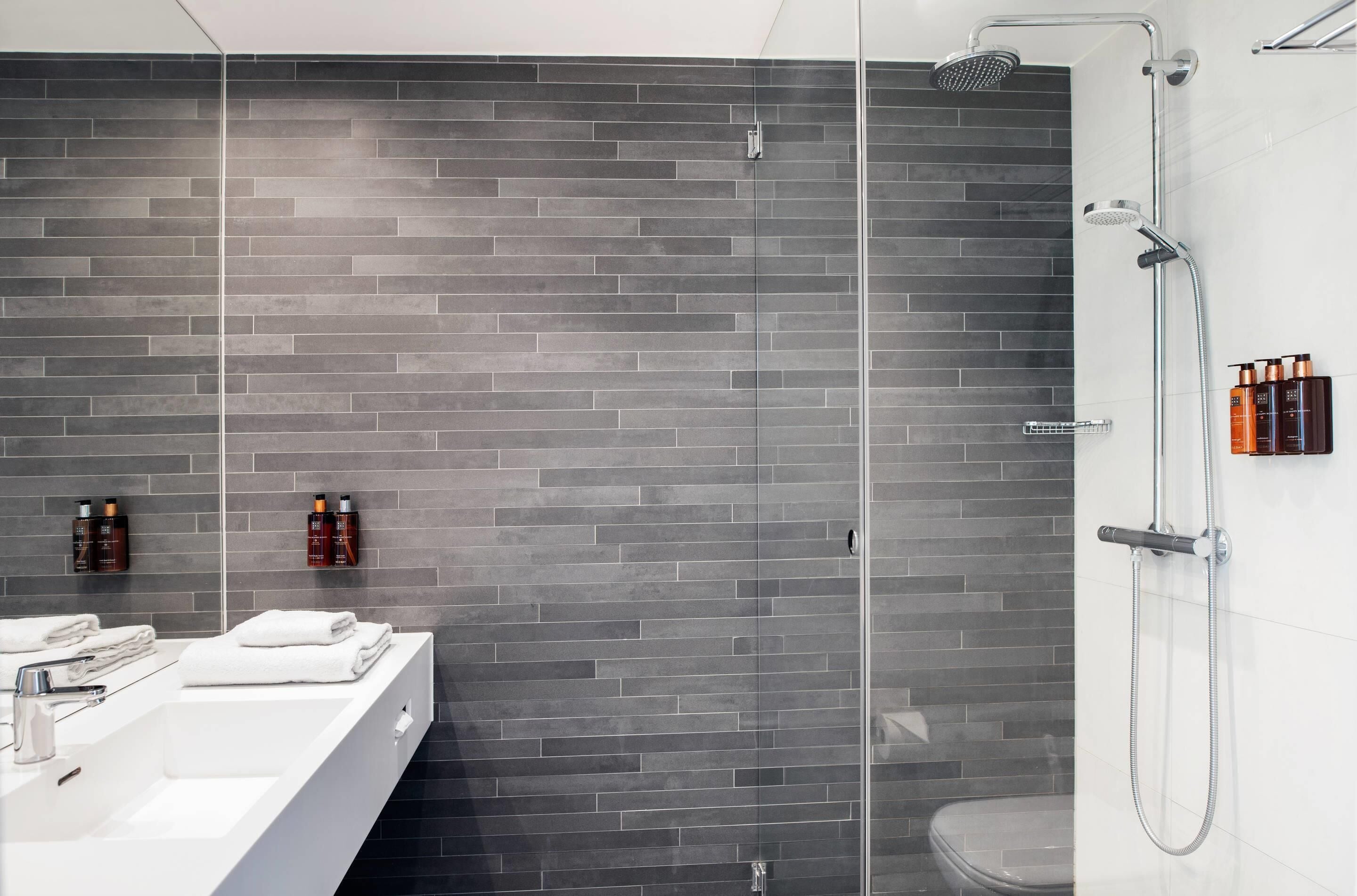 Un baño moderno con pared de ladrillo gris, cabina de ducha de cristal, lavamanos blanco y artículos de aseo colgados en la pared.