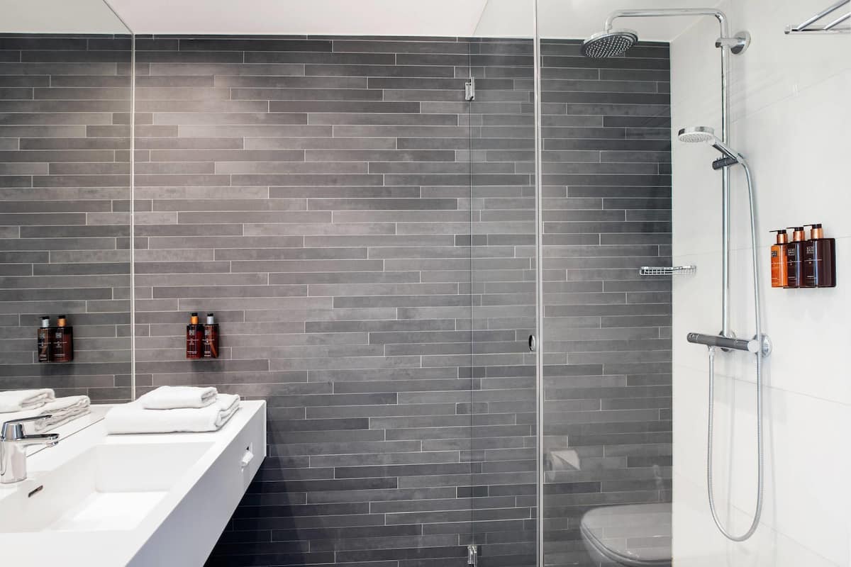 Un baño moderno con pared de ladrillo gris, cabina de ducha de cristal, lavamanos blanco y artículos de aseo colgados en la pared.