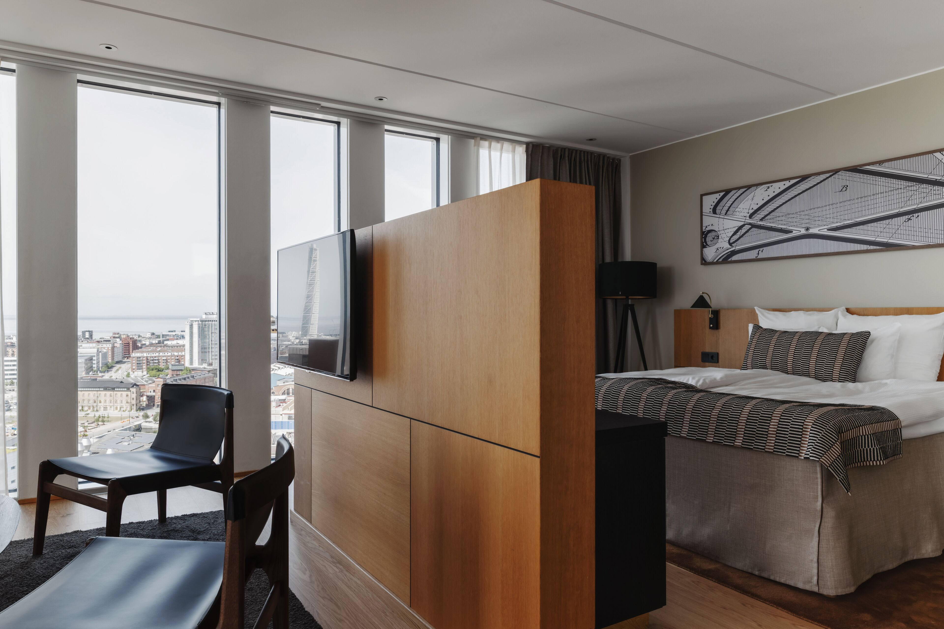 A modern hotel room with a large window, a bed with patterned bedding, a desk with a chair, and a city view.