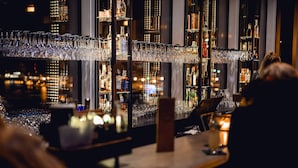 Un bar con numerosas estanterías llenas de botellas y vasos, una barra y vistas a un paisaje urbano a través de amplios ventanales.