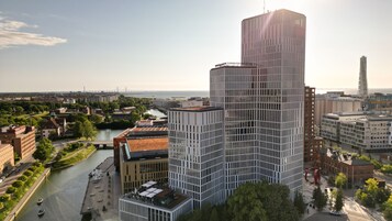 Un edificio moderno de gran altura con fachada de entramado, ubicado en una zona urbana con un río y un puente al fondo.