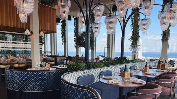 A restaurant with a view of the sea, featuring blue patterned seating, hanging crystal chandeliers, and a bar area.