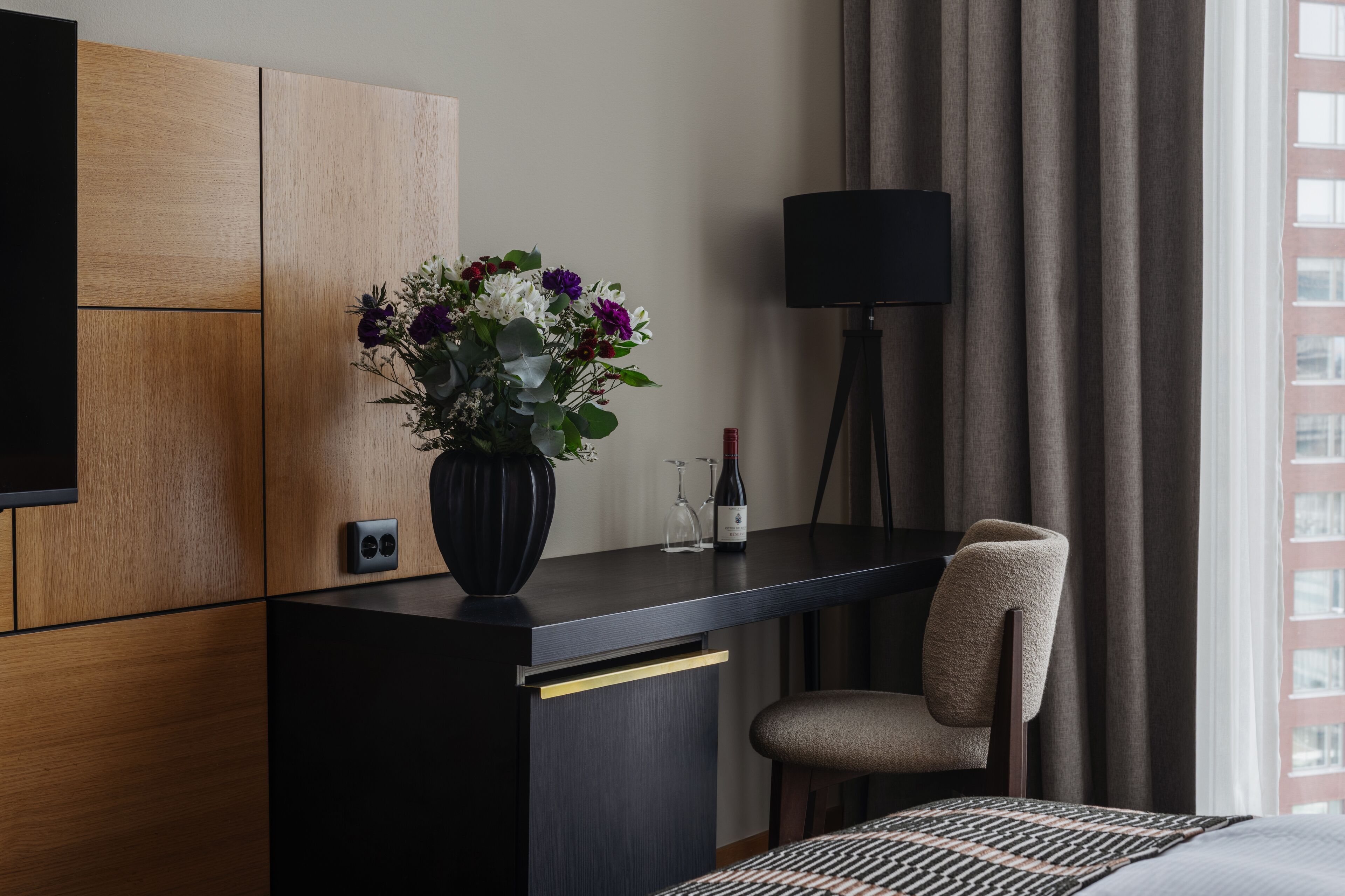 A hotel room with a wooden panel wall, a black desk with a vase of flowers, a wine bottle, and a lamp.