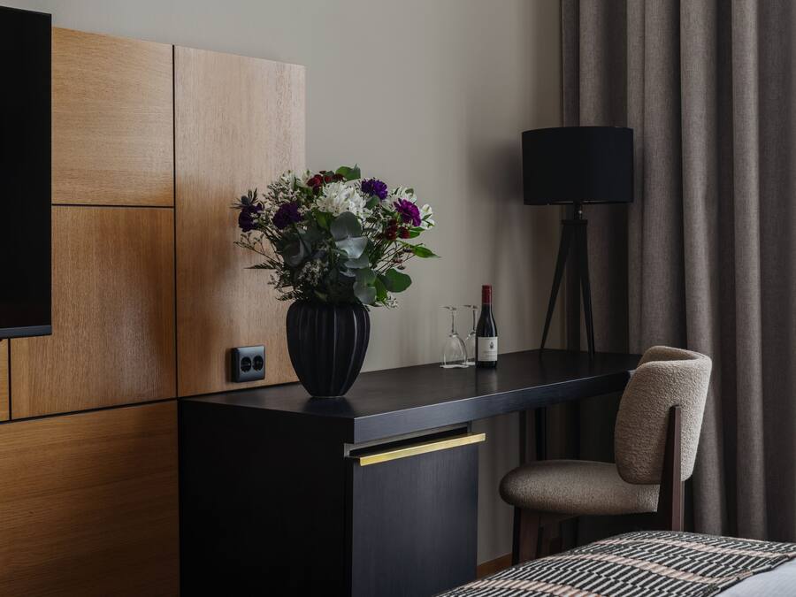 A hotel room with a wooden panel wall, a black desk with a vase of flowers, a wine bottle, and a lamp.