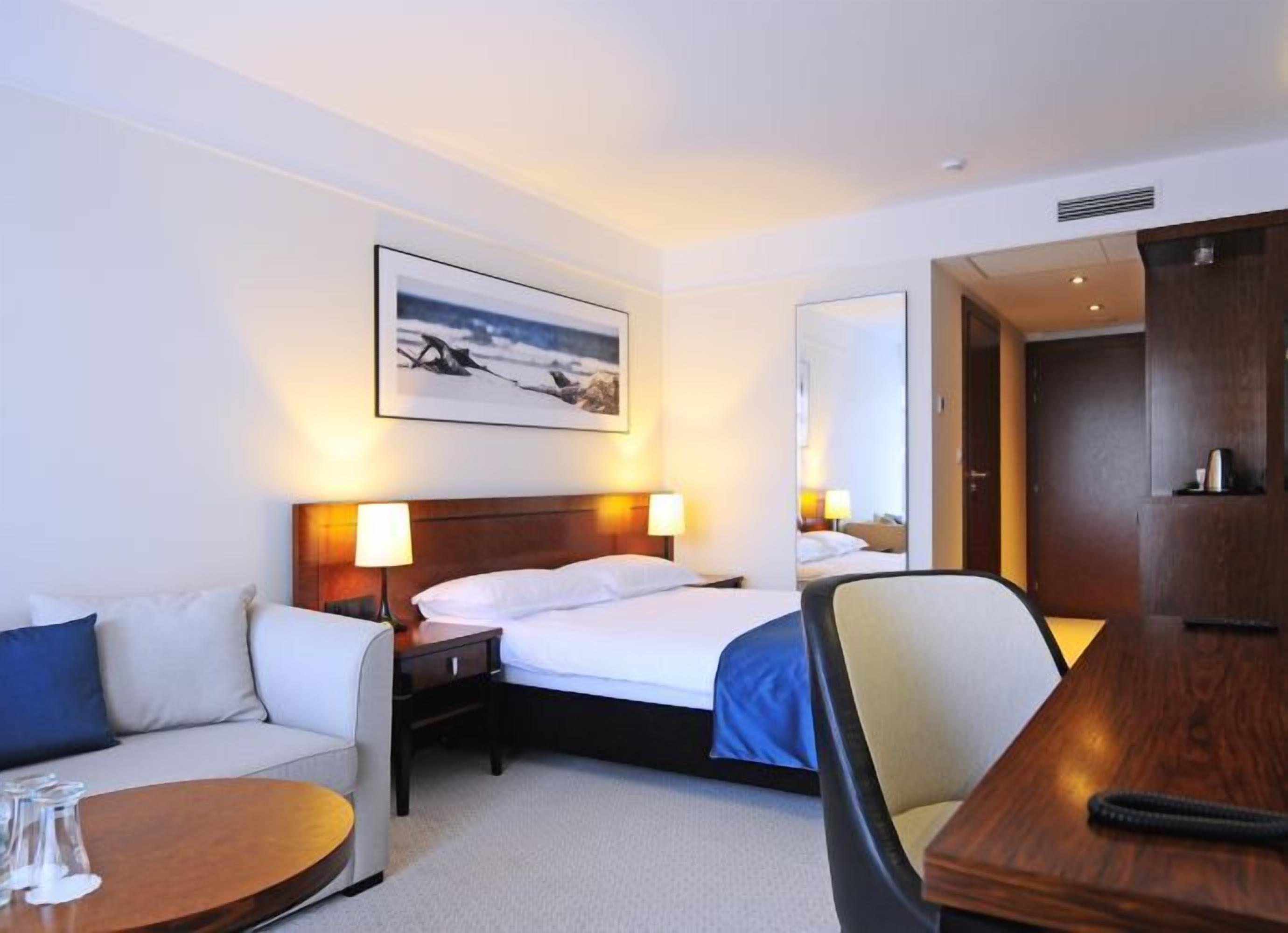 A hotel room with a bed, bedside tables, a sofa, a small round table, and a mirror.