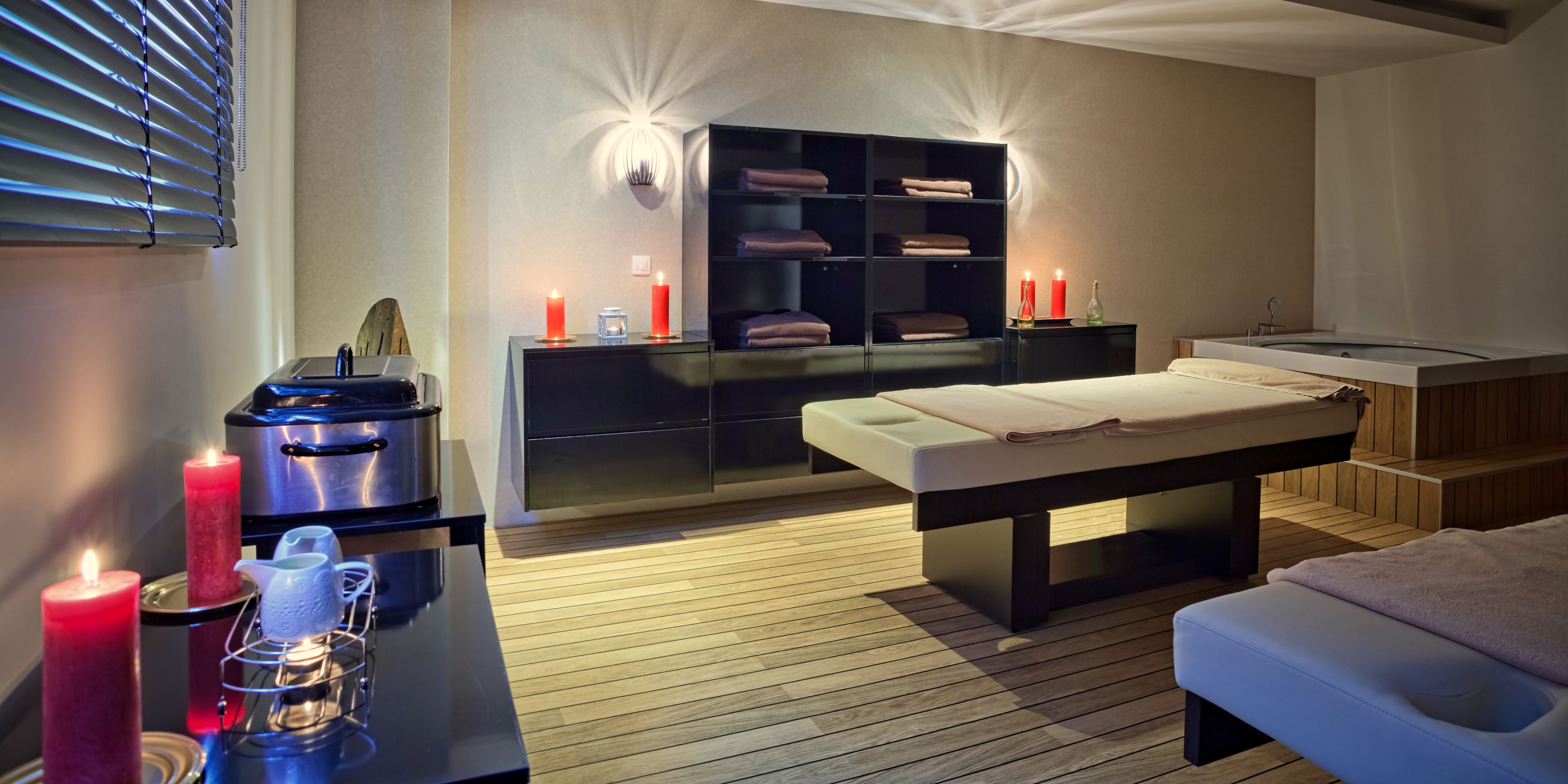 Ein moderner Spa-Raum mit Massageliege, Holzboden, einer schwarzen Regaleinheit mit Handtüchern und Kerzen.