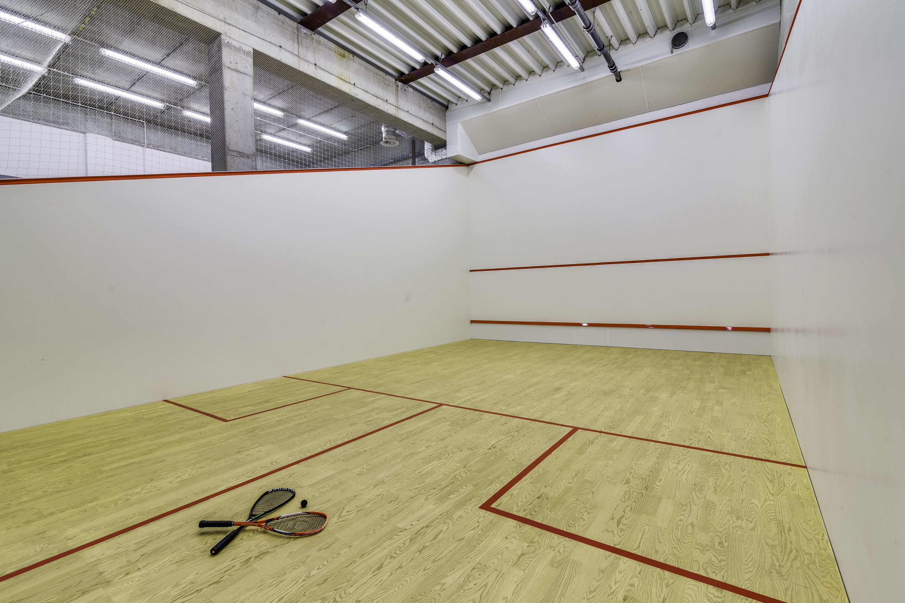 Pista de squash interior con suelo de madera, paredes blancas y líneas rojas que delimitan el campo.