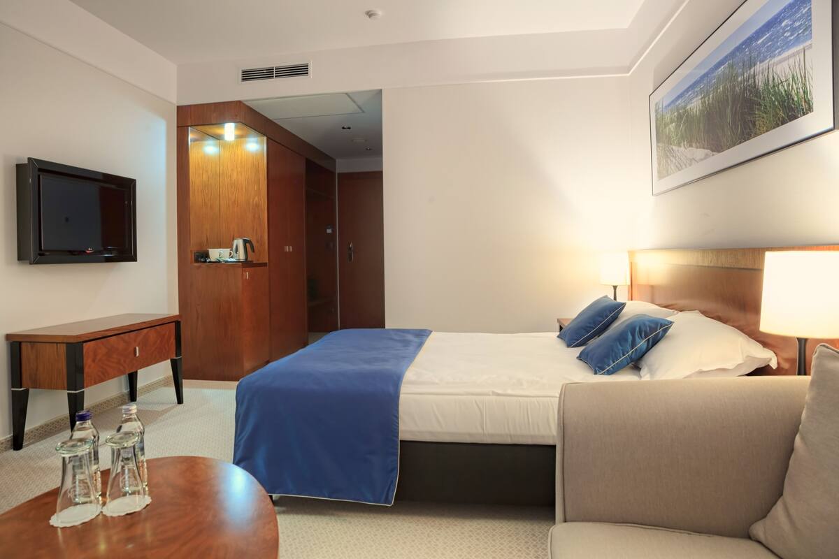 A hotel room with a bed, a desk, a chair, a TV, and a painting on the wall.