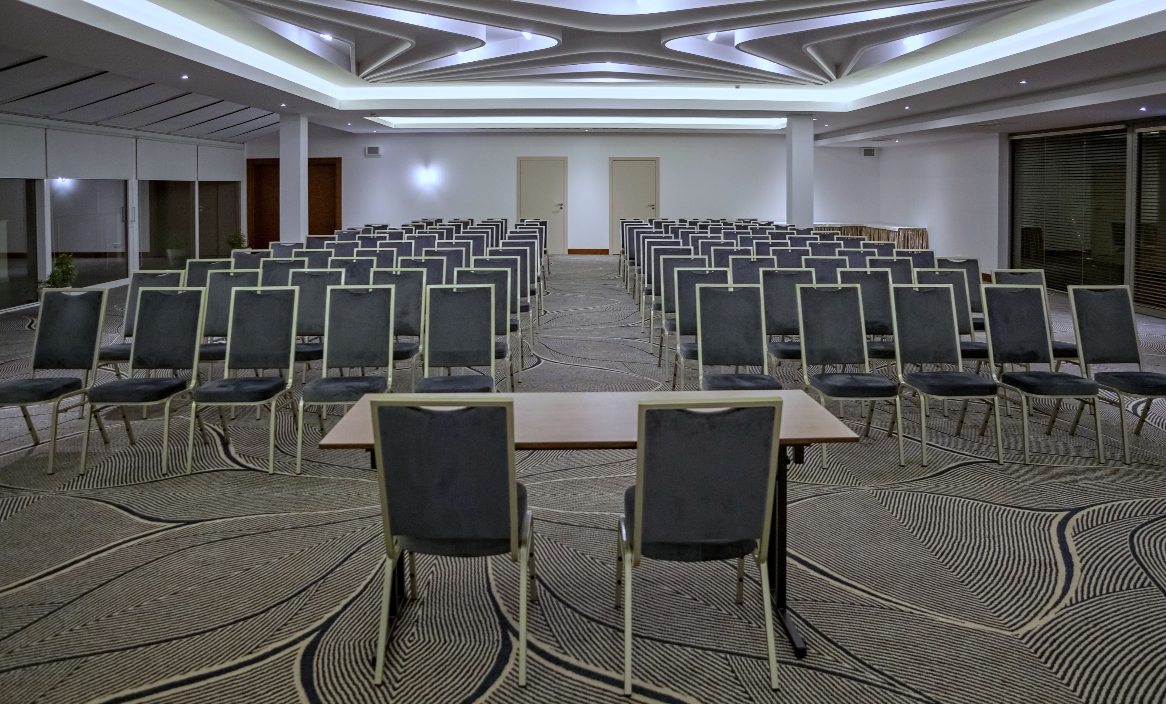 A conference room with rows of chairs and a long table, carpeted floor, and modern ceiling design.