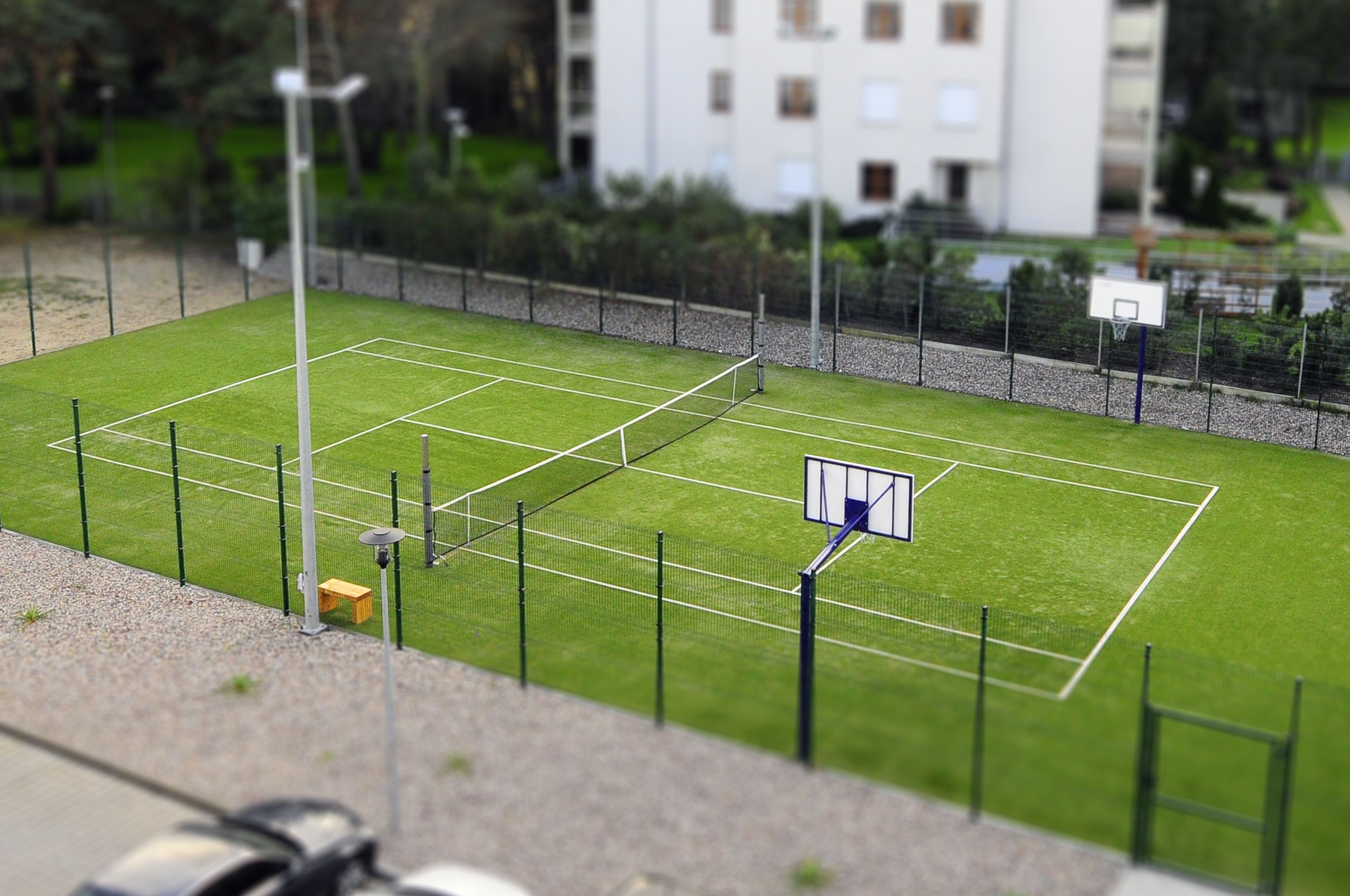 A tennis court with a net, surrounded by a fence, and a basketball hoop.