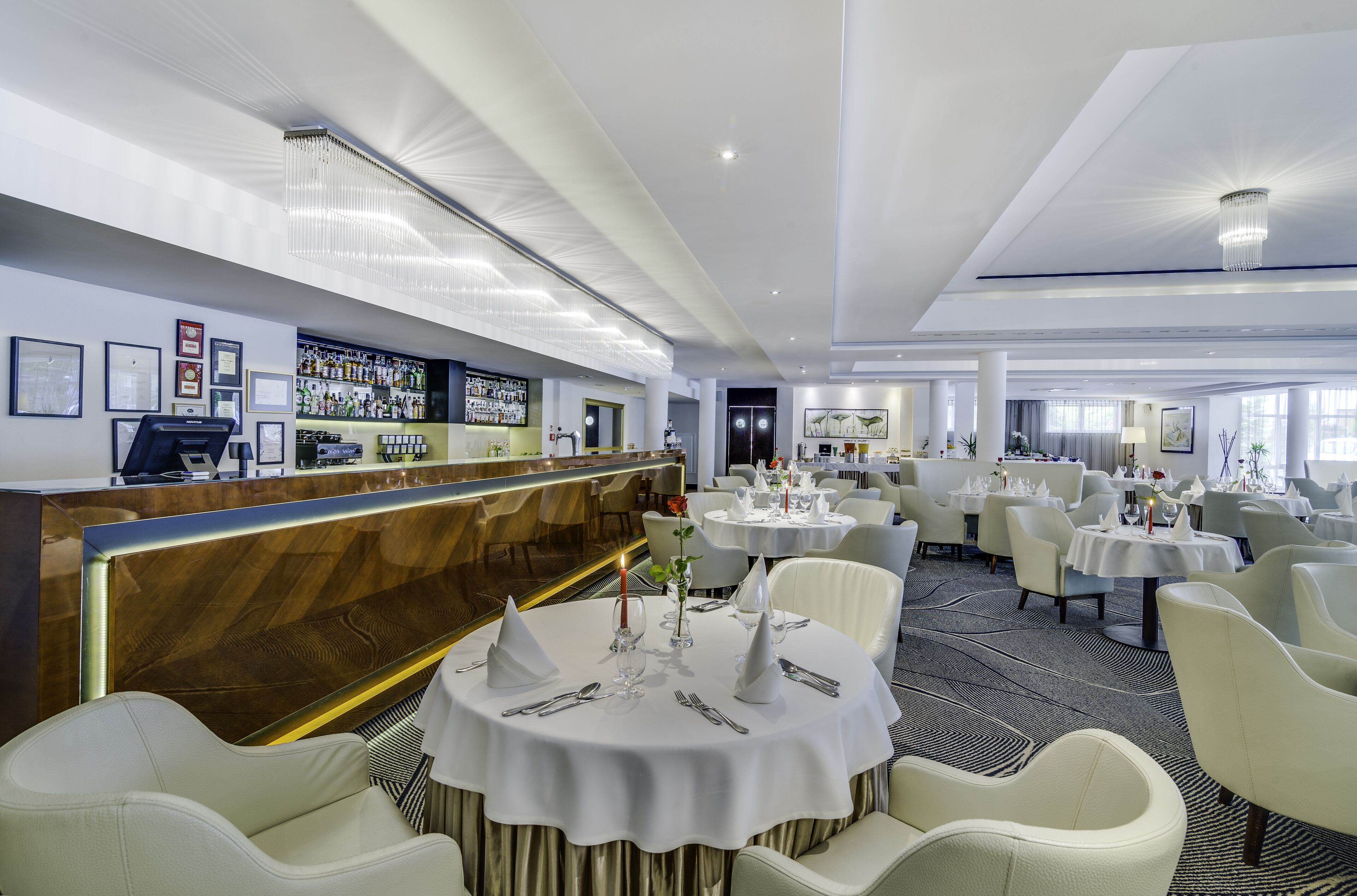 Un restaurant avec des tables rondes, des chaises blanches, un bar et des tableaux encadrés aux murs.