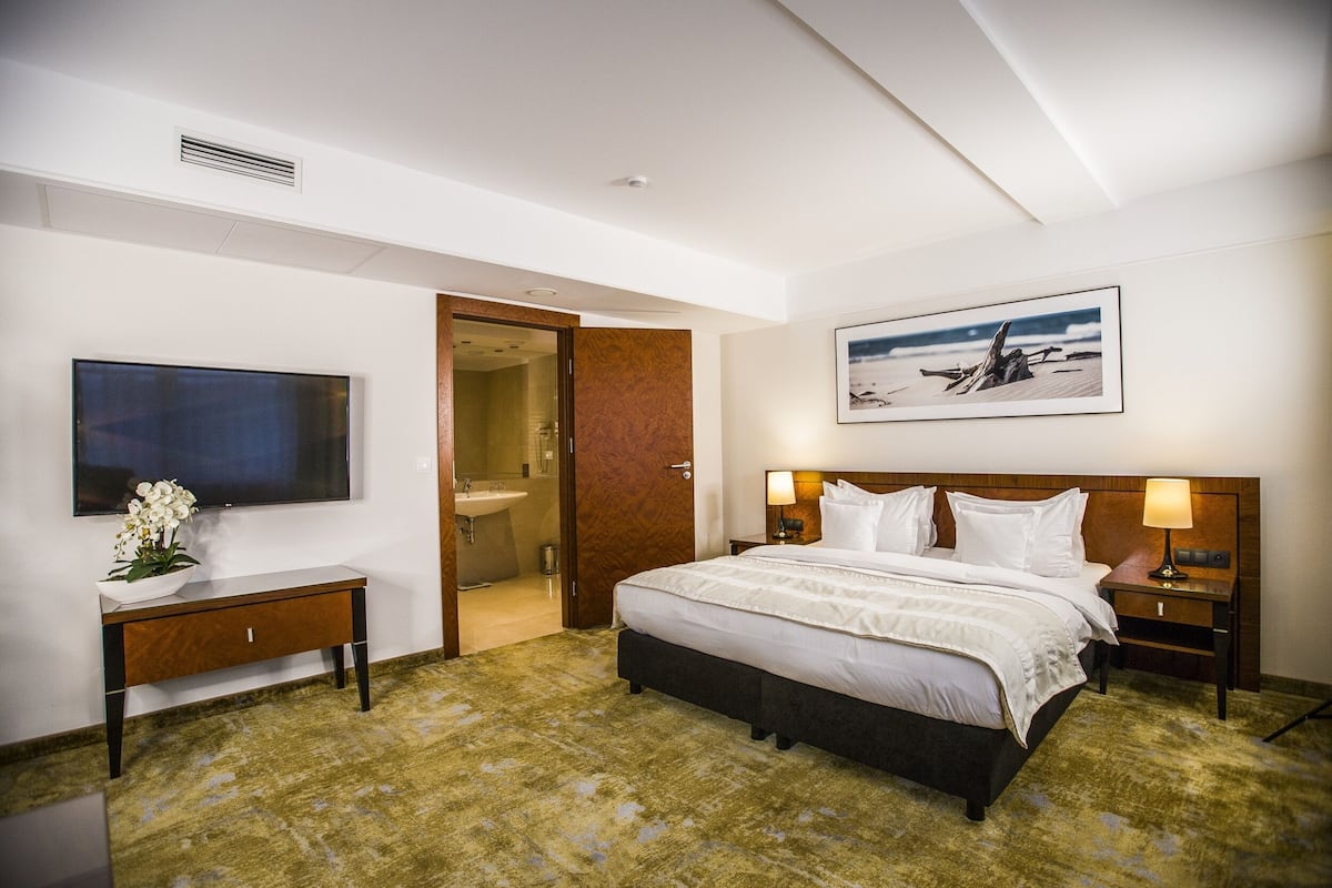 A hotel room with a large bed, a flat-screen TV, a wooden bedside table, a nightstand, and a framed picture on the wall.