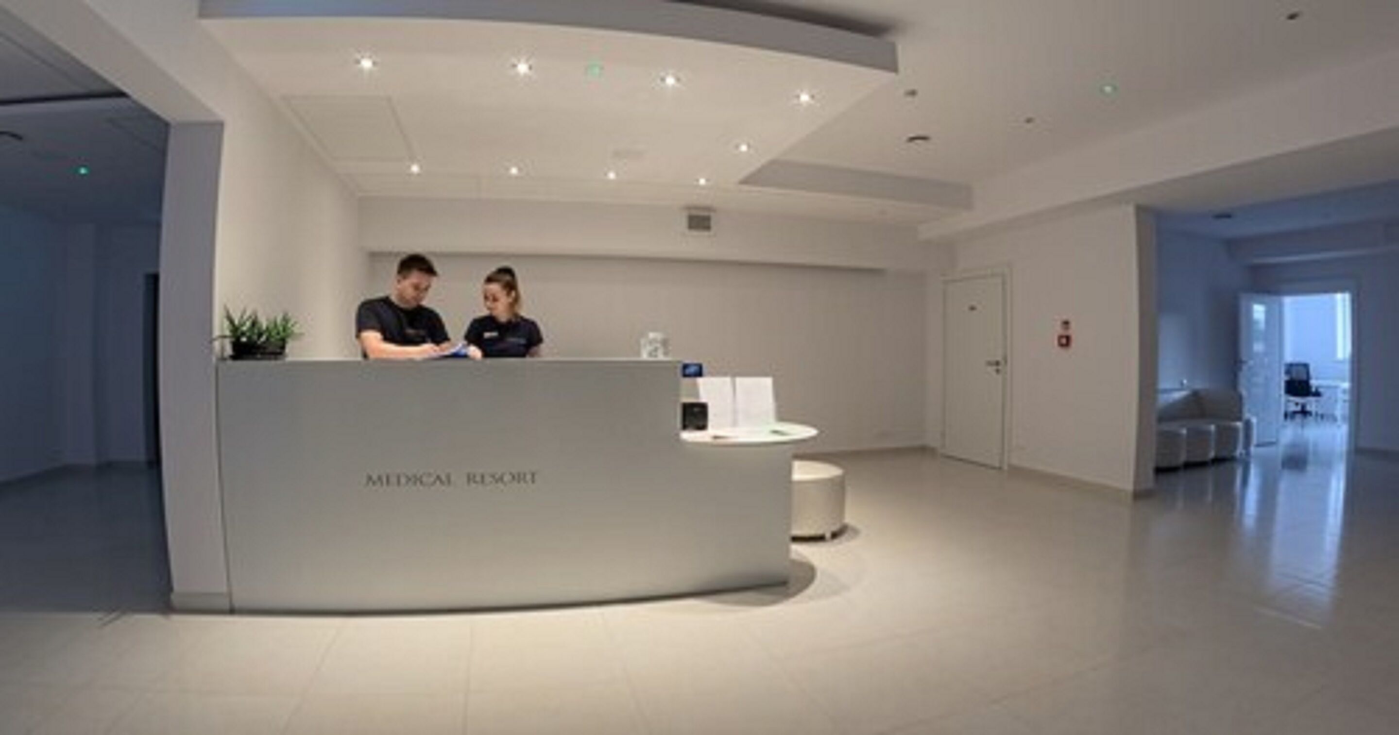 A reception desk labeled "MEDICAL RESORT" with two staff members, a round white table, and a plant.