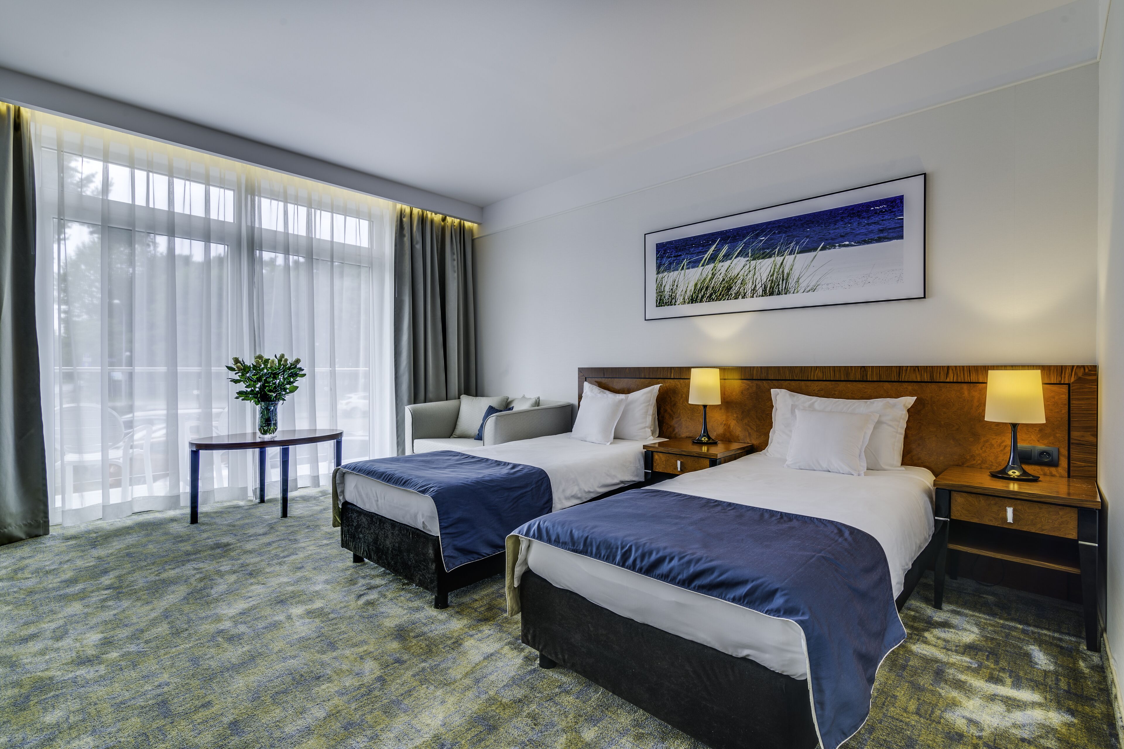 A hotel room with two beds, a small table with a plant, a nightstand with a lamp, and a large window with curtains.