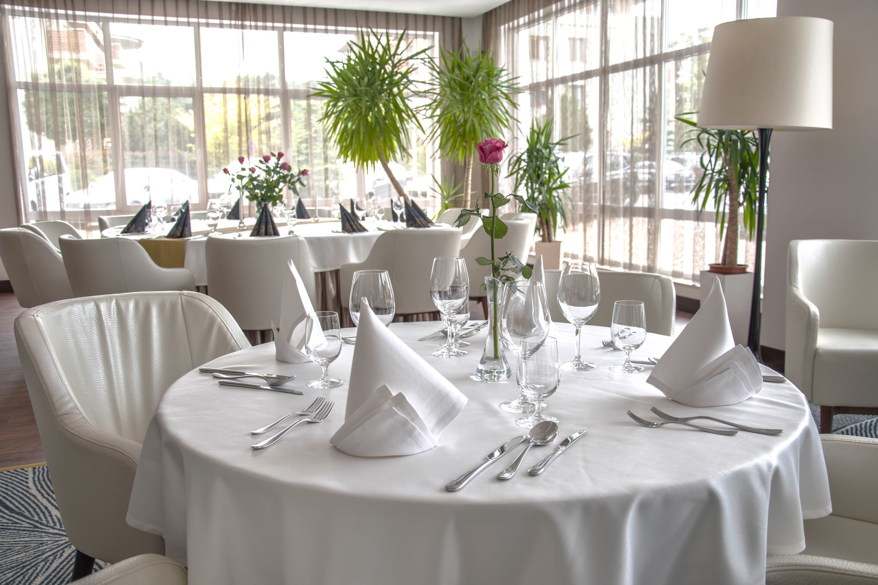 Un comedor con mesas redondas dispuestas para una comida, con manteles blancos, servilletas dobladas y cubiertos.