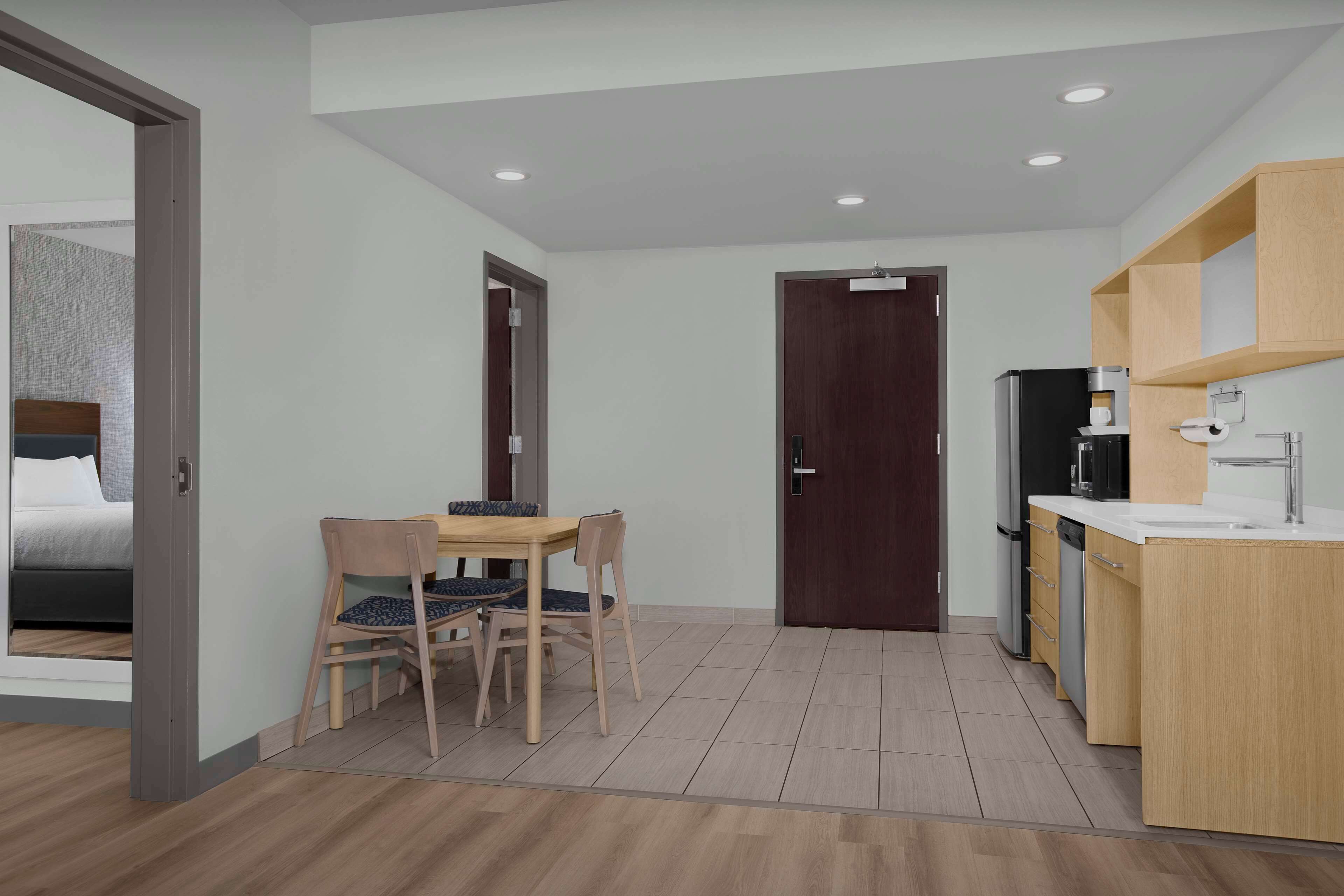 Una habitación de hotel moderna con una mesa y sillas de madera, una pequeña zona de cocina con un refrigerador y cafetera, y una puerta que da a otra habitación.