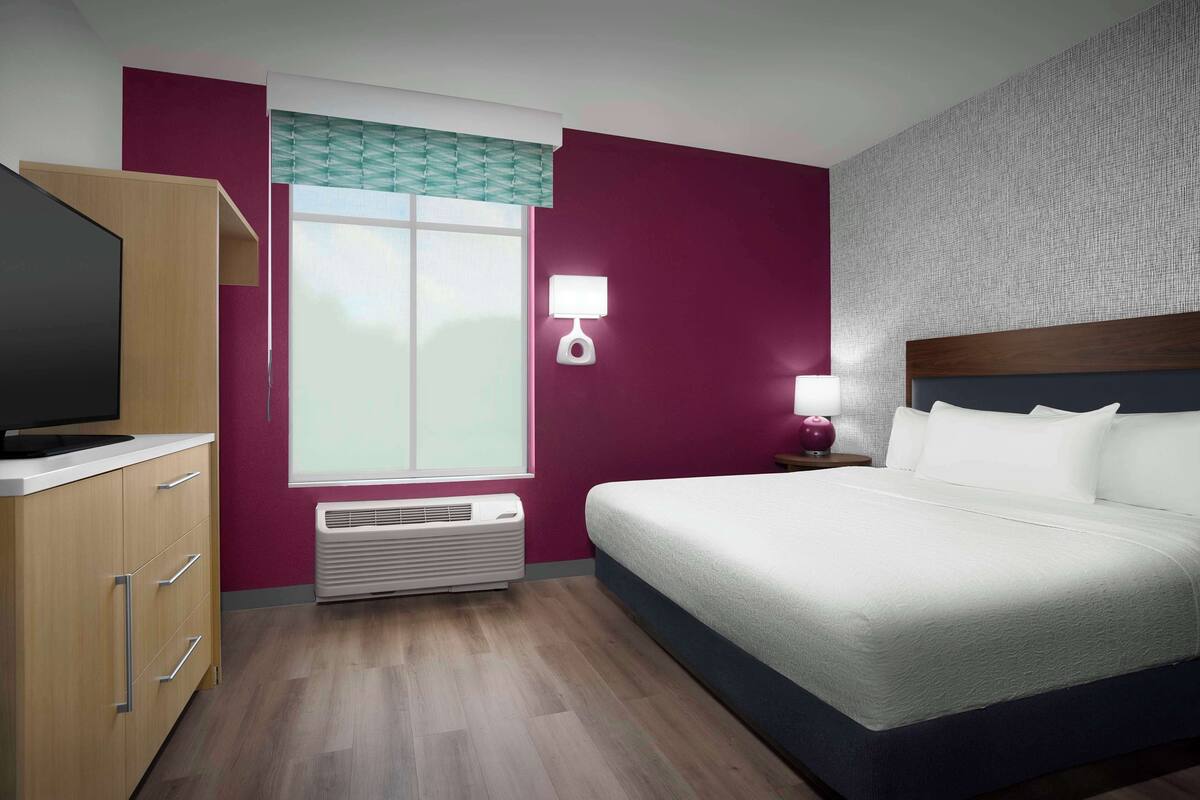 Ein Hotelzimmer mit Bett, Fernseher, Nachttisch mit Lampe, Fenster mit Aussicht und Wandheizung.