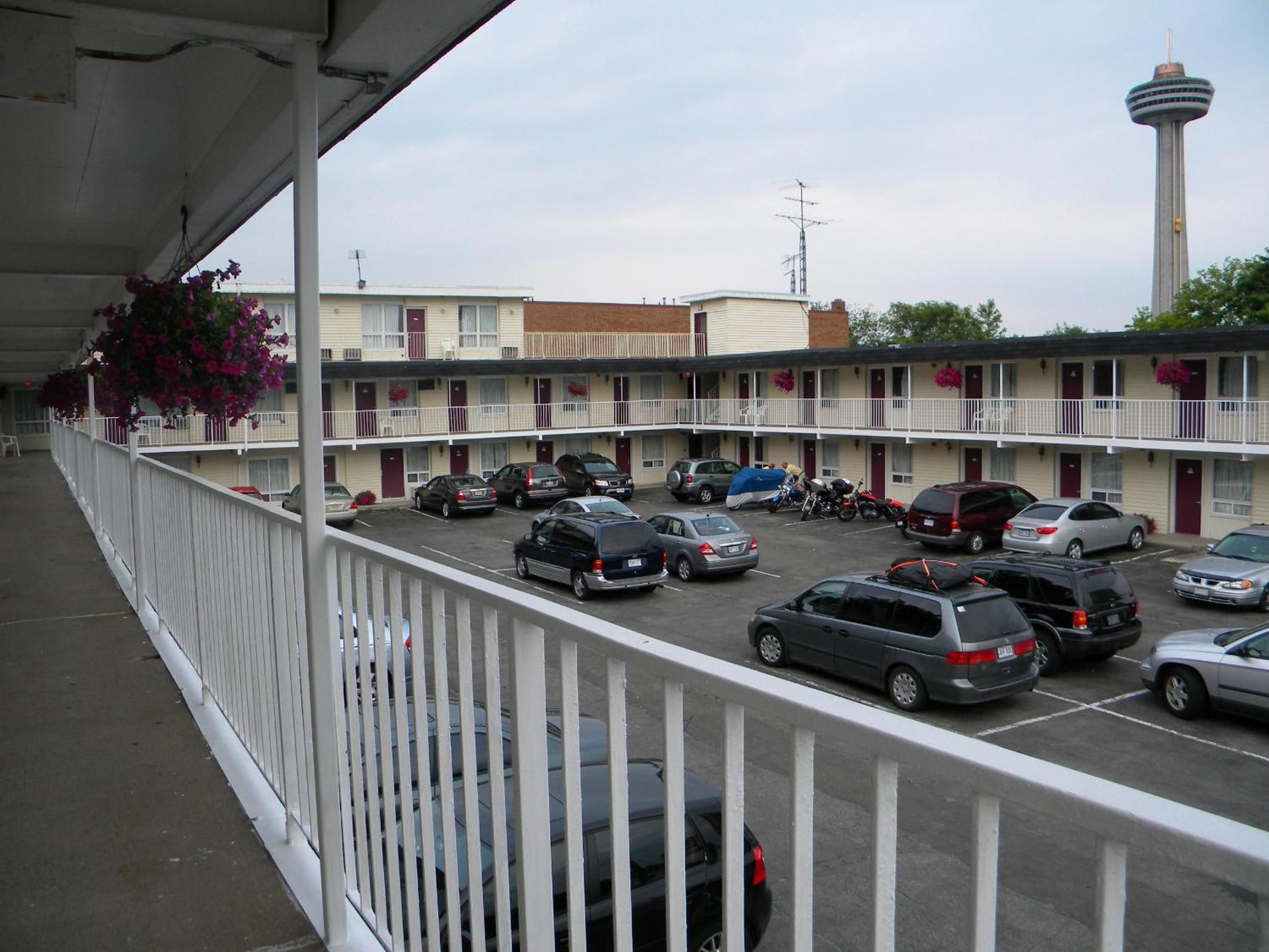 A motel parking lot with multiple cars parked, a tower in the background, and a balcony with a railing.