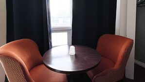 Quarto com duas cadeiras laranja em torno de uma mesa pequena, uma janela com cortinas e uma porta com cabideiro.