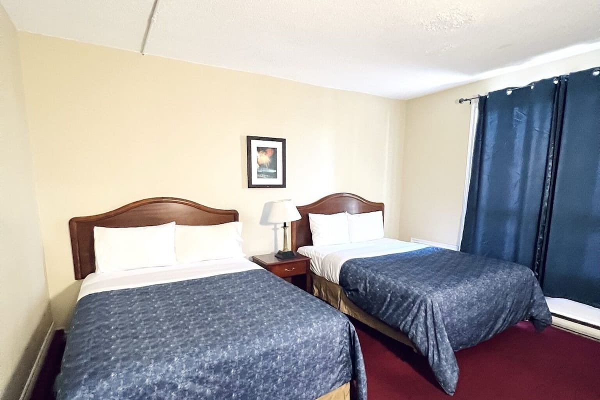A hotel room with two beds, a nightstand, a lamp, a framed picture on the wall, and a window with curtains.