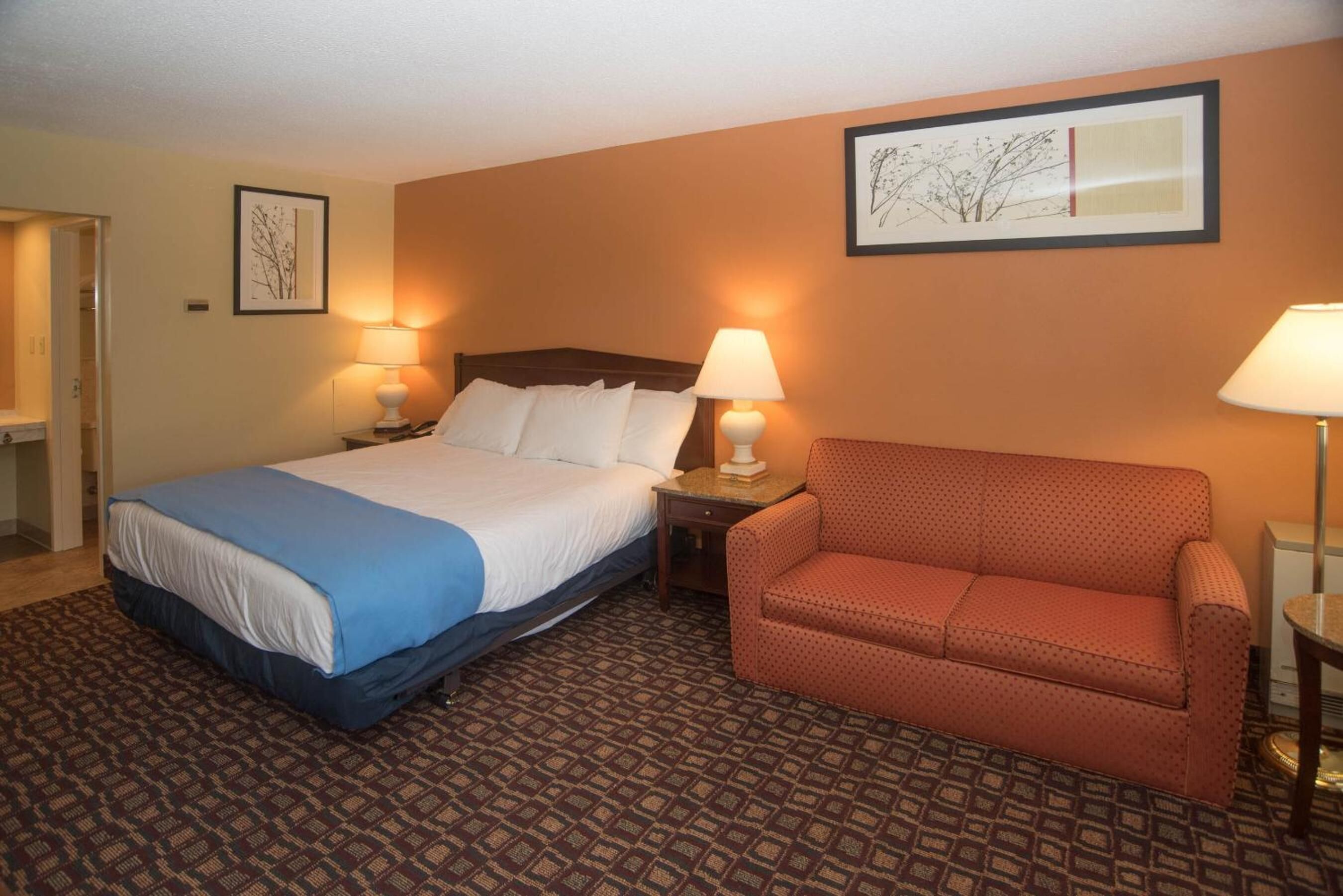 A hotel room with a bed, a sofa, two lamps, and framed artwork on the walls.