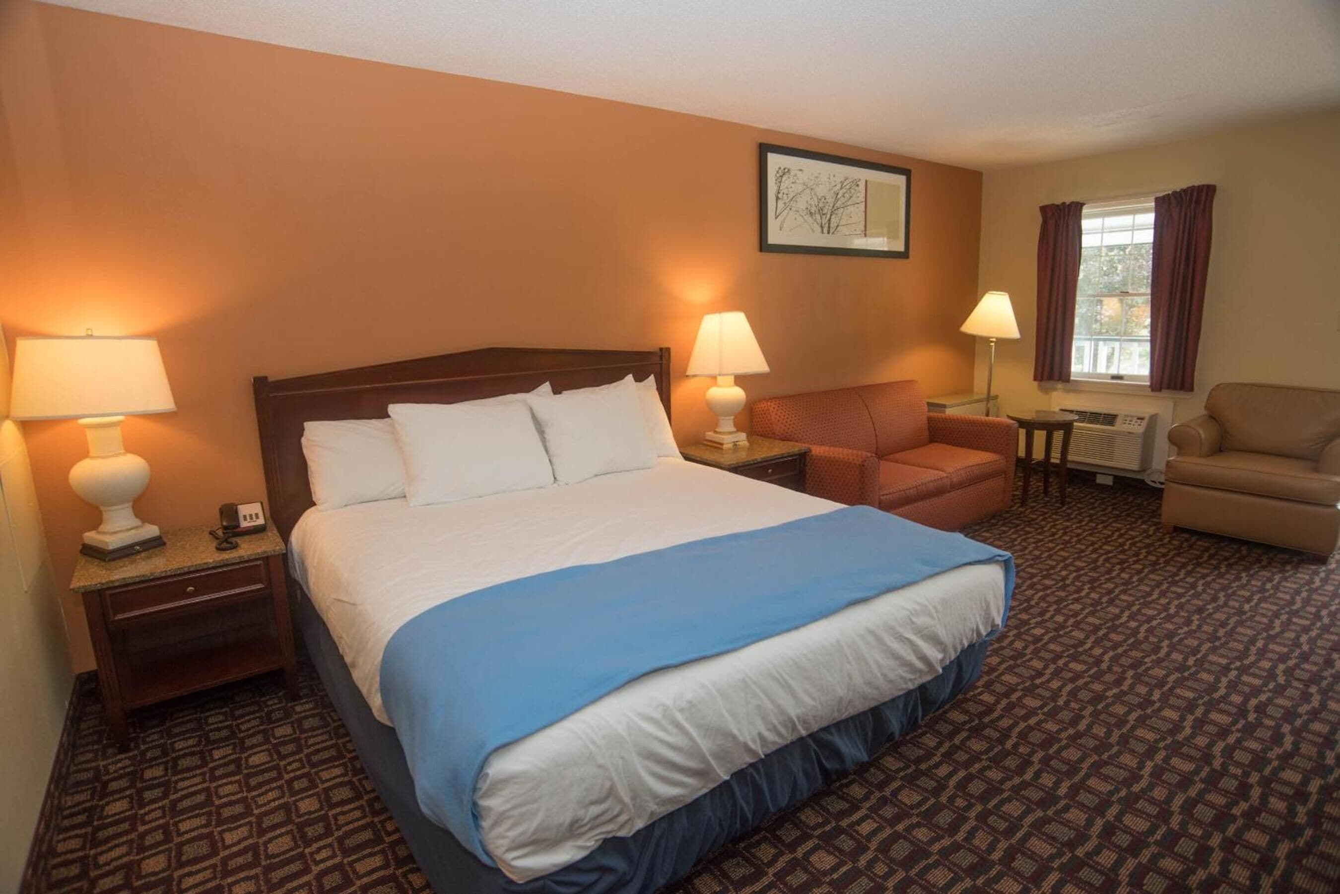 Een hotelkamer met een groot bed, twee nachtkastjes met lampen, een stoel, een klein tafeltje en een raam met gordijnen.