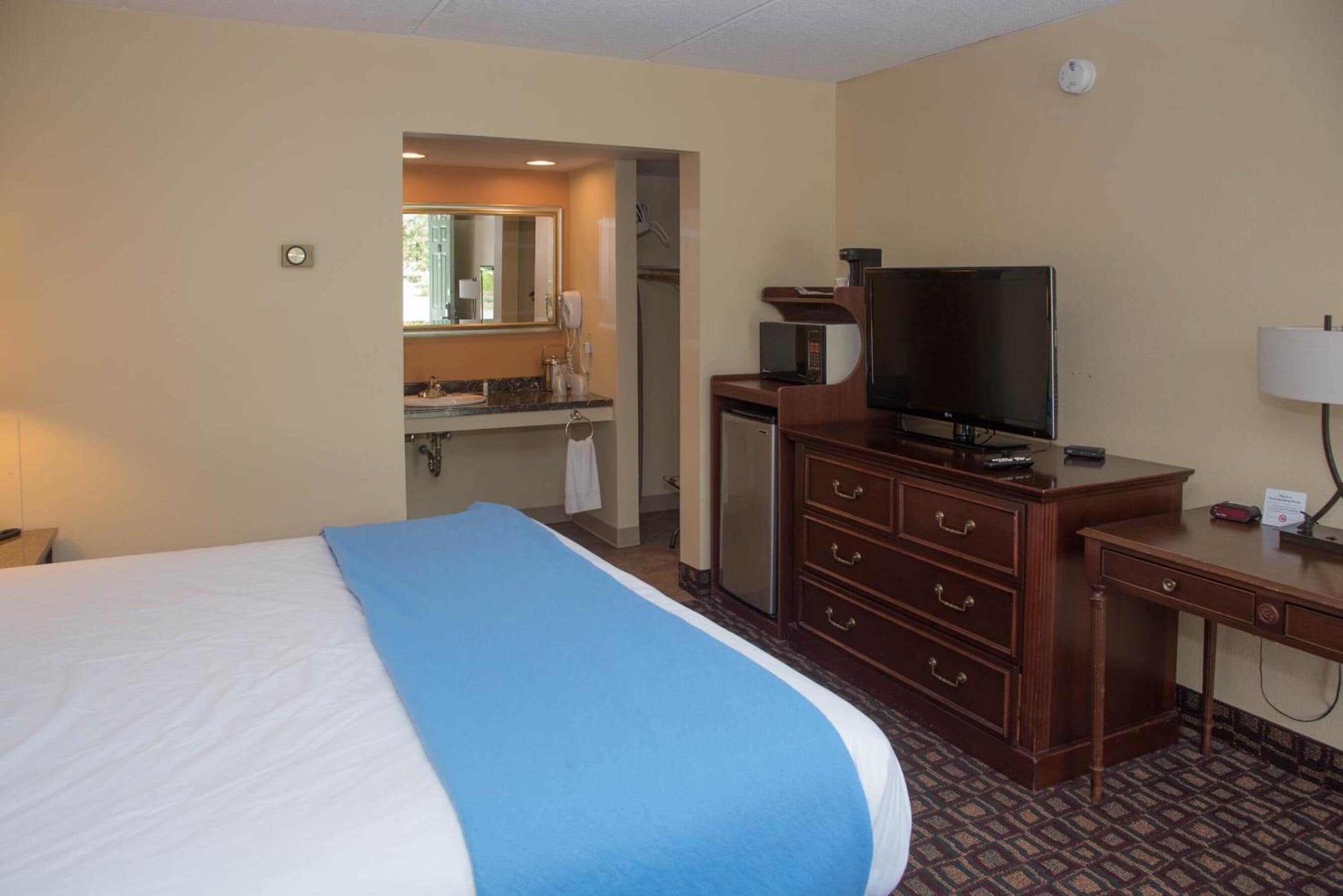 Habitación de hotel con cama, escritorio, televisor, microondas y baño visible a través de una puerta abierta.