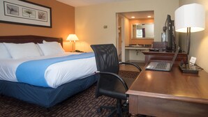 A hotel room with a large bed, a desk with a laptop, a chair, a lamp, and a bathroom visible through an open door.