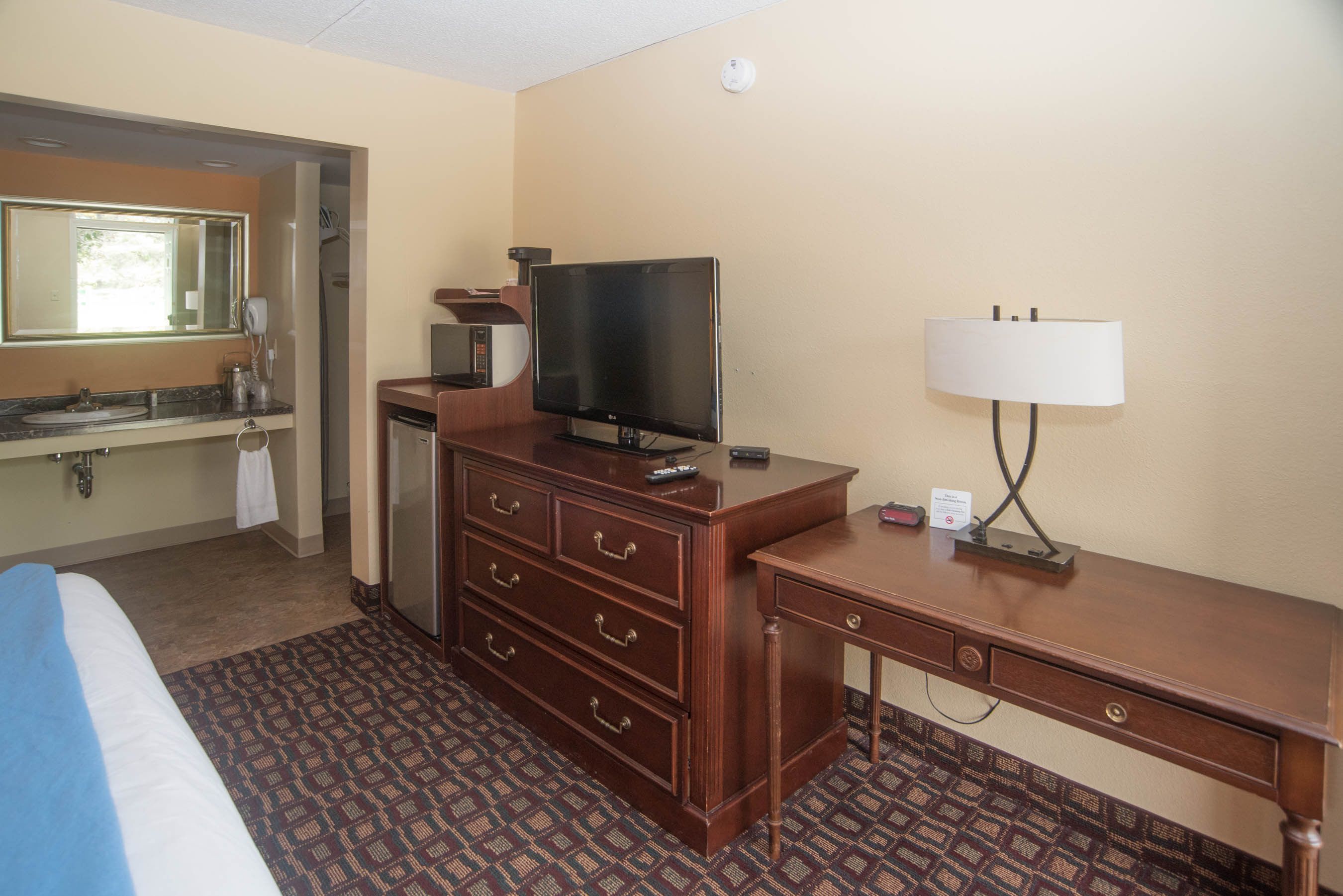 A hotel room with a bed, dresser, television, desk, lamp, and a bathroom with a sink and mirror.