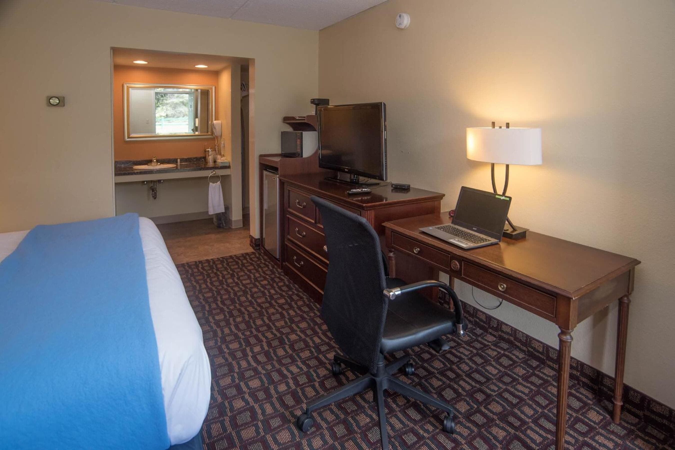 A hotel room with a bed, desk, chair, computer, TV, and a bathroom with a mirror and sink.