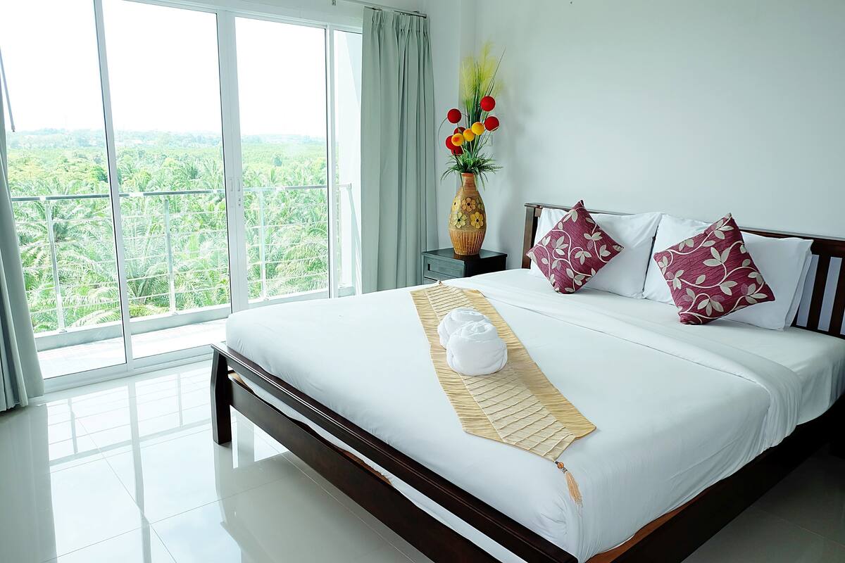 Una cama bien hecha con sábanas blancas, un jarrón decorativo con flores y vistas a exuberante follaje a través de amplios ventanales.