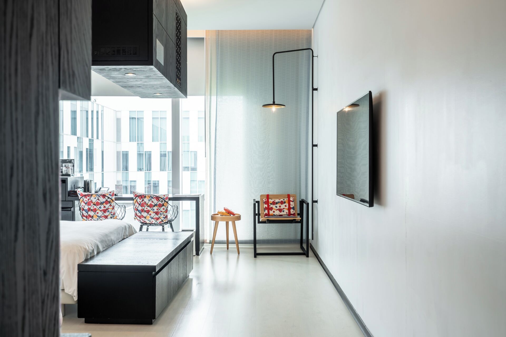 A modern, well-lit room with a large window, a bench, two chairs, a small table, and a wall-mounted TV.