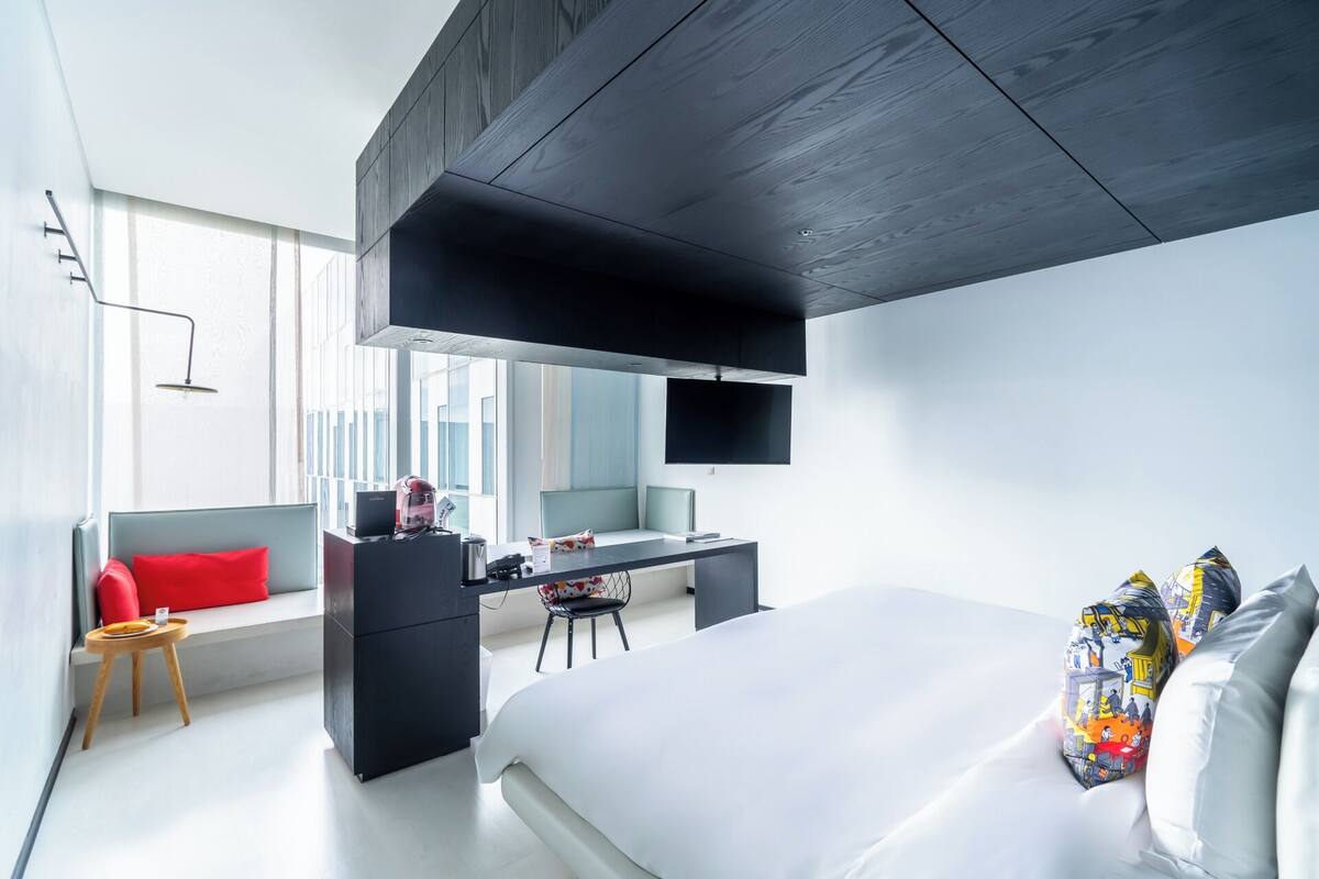 A modern hotel room with a bed, a desk, a chair, and a large window.