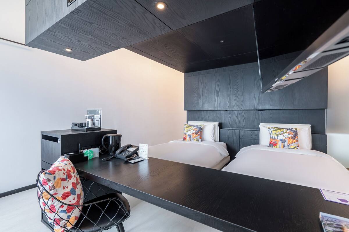 Ein modernes Hotelzimmer mit einem dunklen Holz-Kopfteil, einem Schreibtisch mit Kaffeemaschine, Telefon und einem bunten Stuhl.