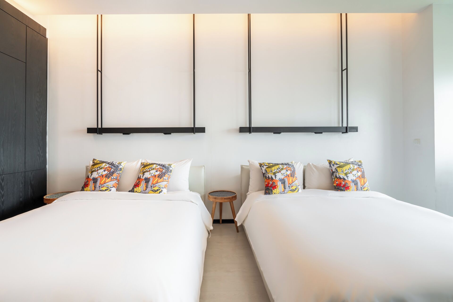 A hotel room with two beds, each with colorful patterned pillows, a small wooden side table, and a round mirror.