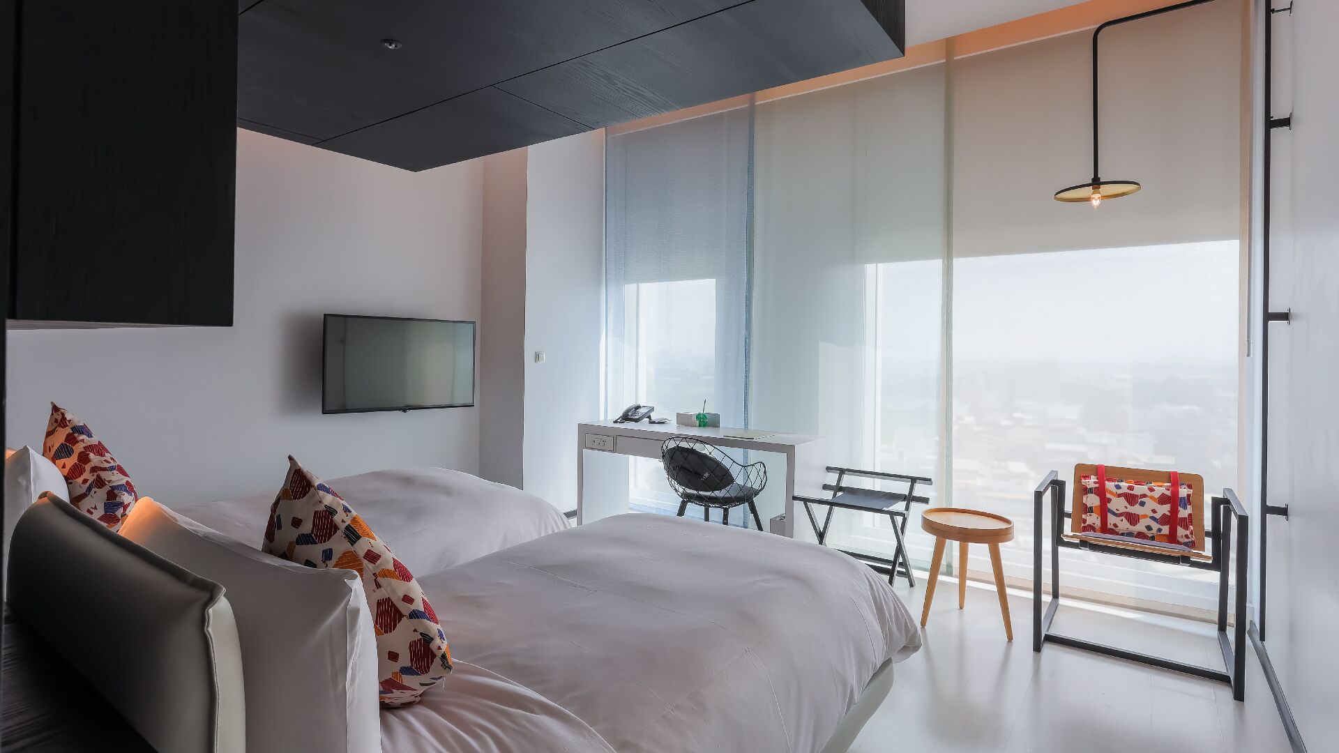 Une chambre d’hôtel moderne, dotée d’un grand lit, d’un bureau et d’une chaise. Un téléviseur est fixé au mur et une fenêtre offre une vue sur la ville.