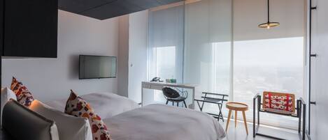 Une chambre d’hôtel moderne, dotée d’un grand lit, d’un bureau et d’une chaise. Un téléviseur est fixé au mur et une fenêtre offre une vue sur la ville.