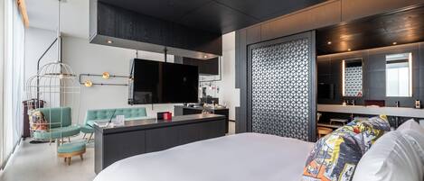 A modern hotel room with a large bed, a desk area, a TV, and a bathroom with a mirror.