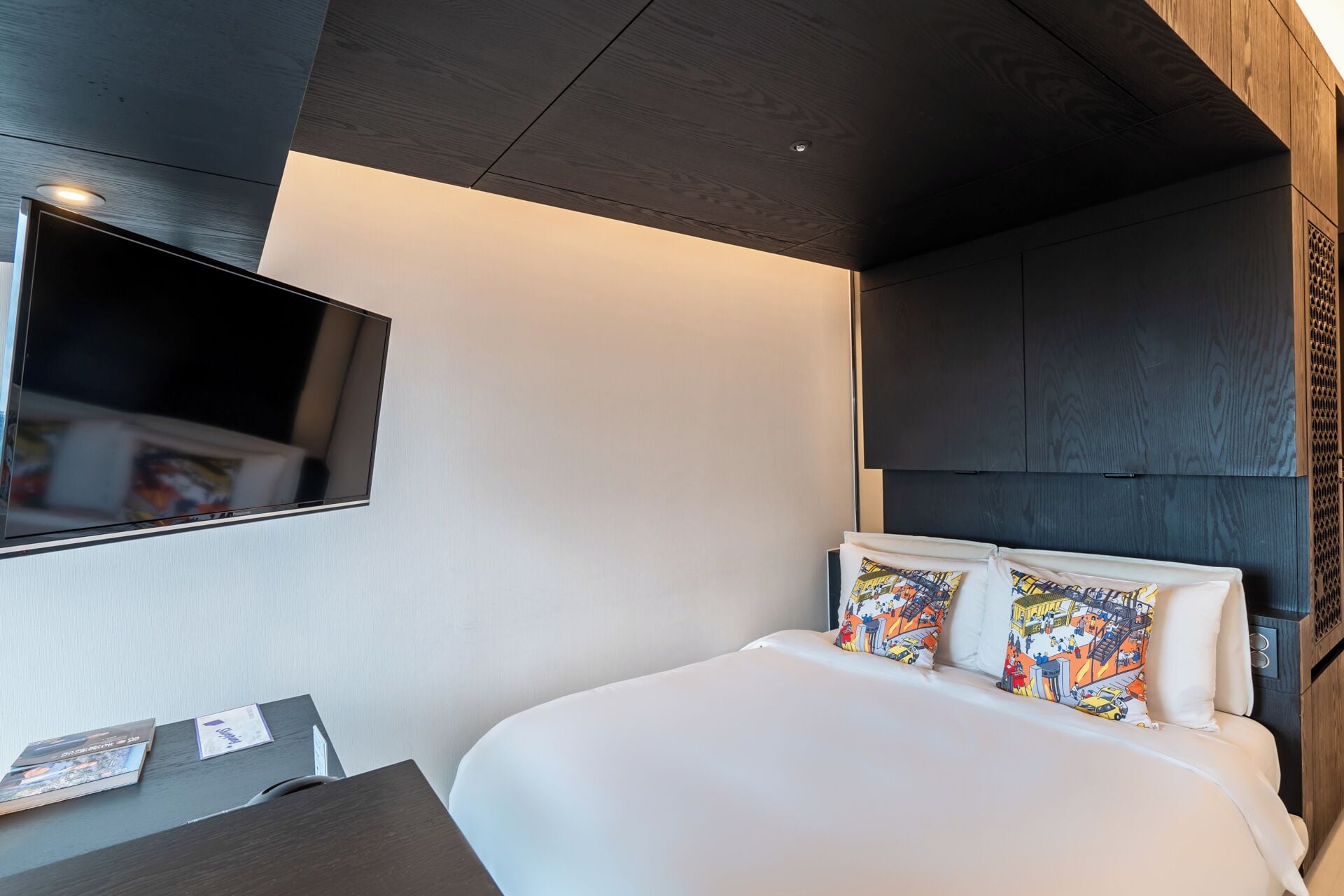 A modern hotel room with a large bed, a flat-screen TV mounted on the wall, and a dark wooden headboard.