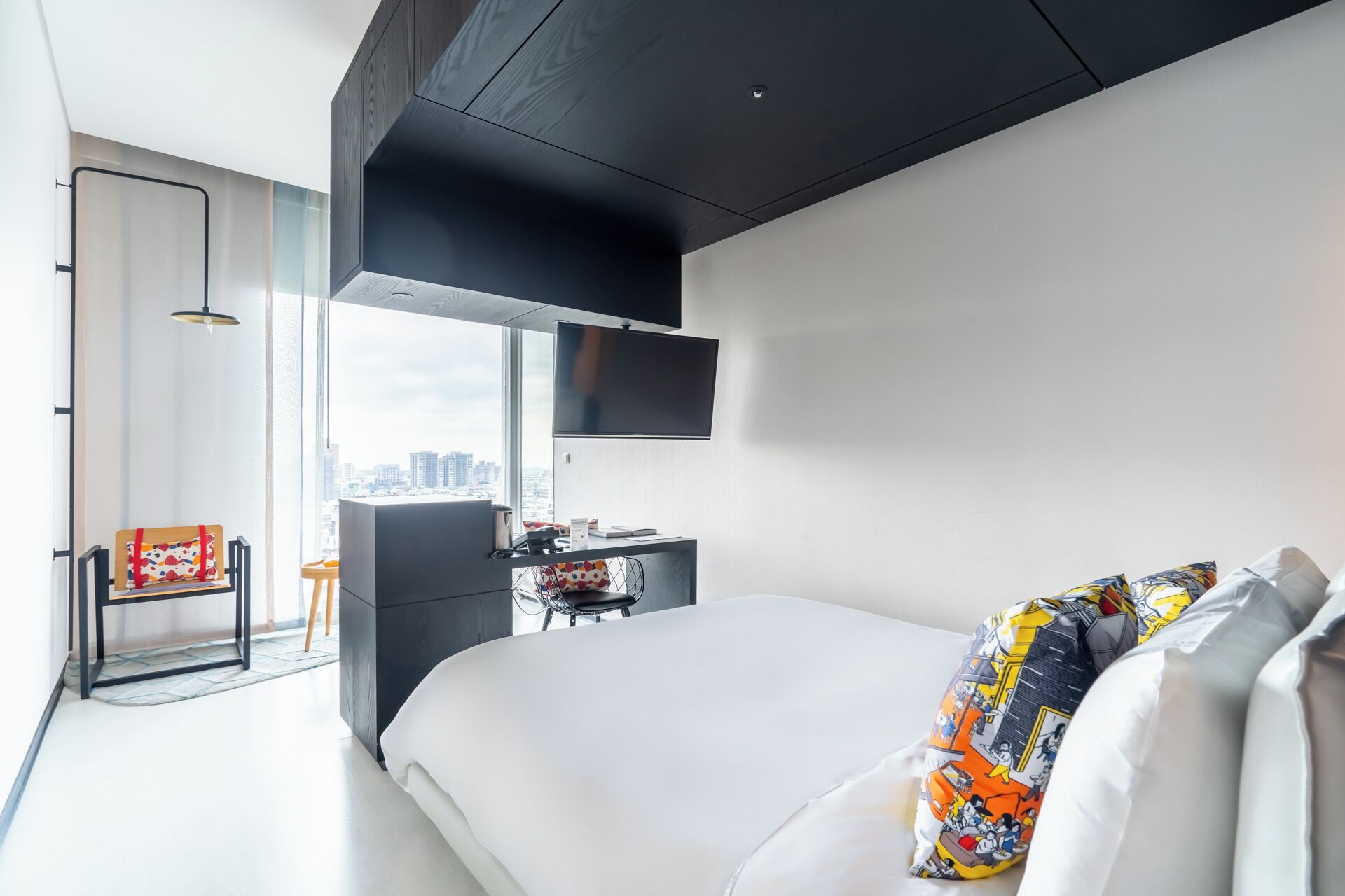 A modern hotel room with a large bed, a desk with a chair, a TV mounted on the wall, and a city view through the window.