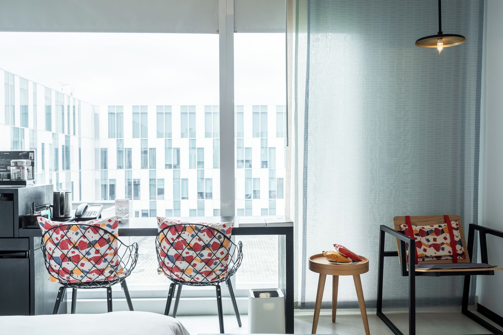 A modern hotel room with a large window, two patterned chairs, a small table with fruit, and a hanging light fixture.