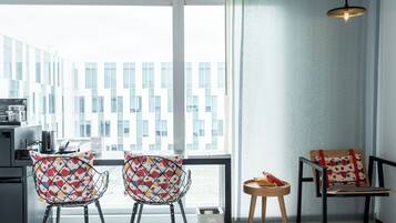 A modern hotel room with a large window, two patterned chairs, a small table with fruit, and a hanging light fixture.