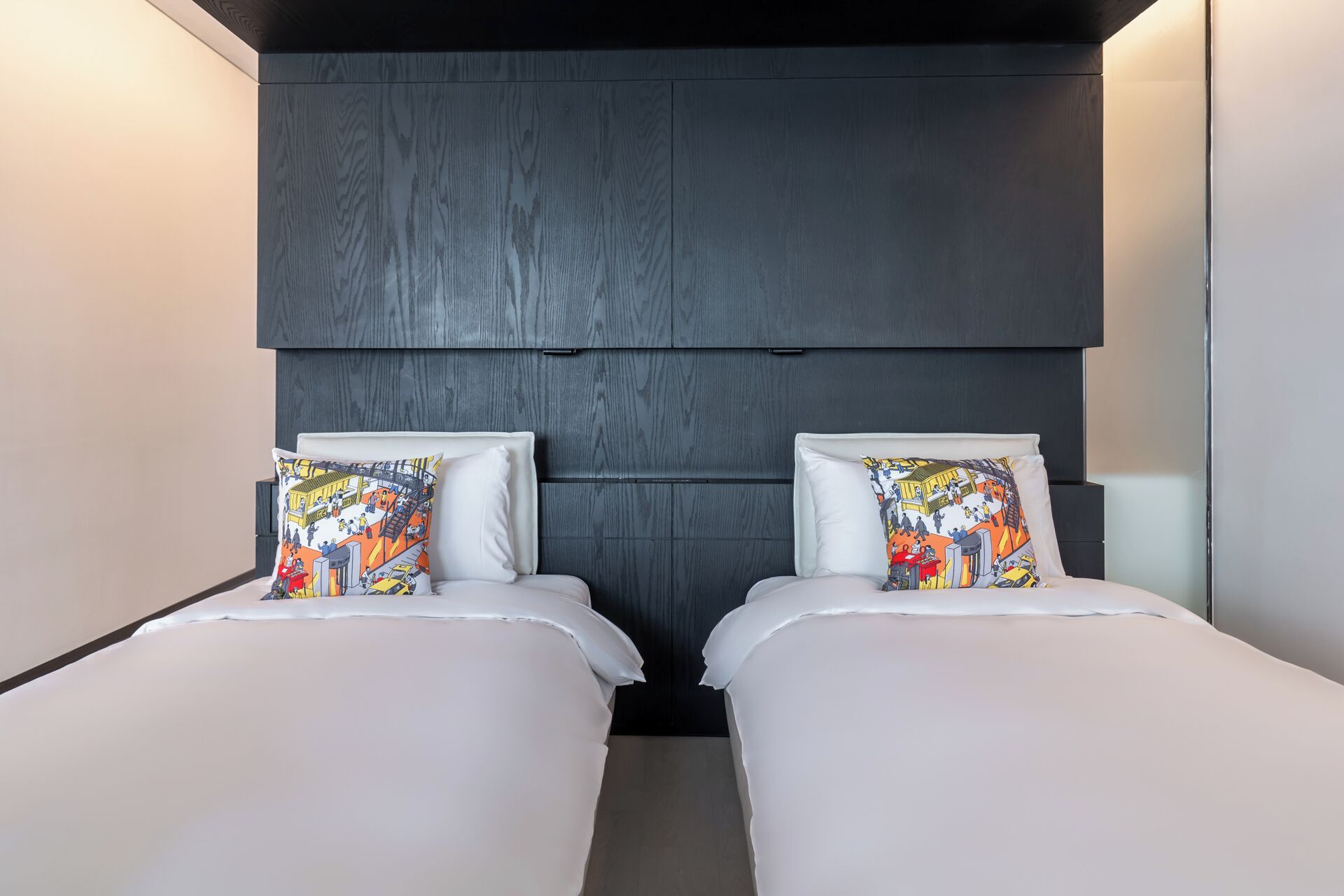 A room with two beds, each with a colorful pillow and white bedding. The room has a dark wood panel headboard and a light-colored wall.