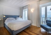 A bedroom with a large bed, a blue headboard, a nightstand with a lamp, a blue chair, and a balcony with a view.
