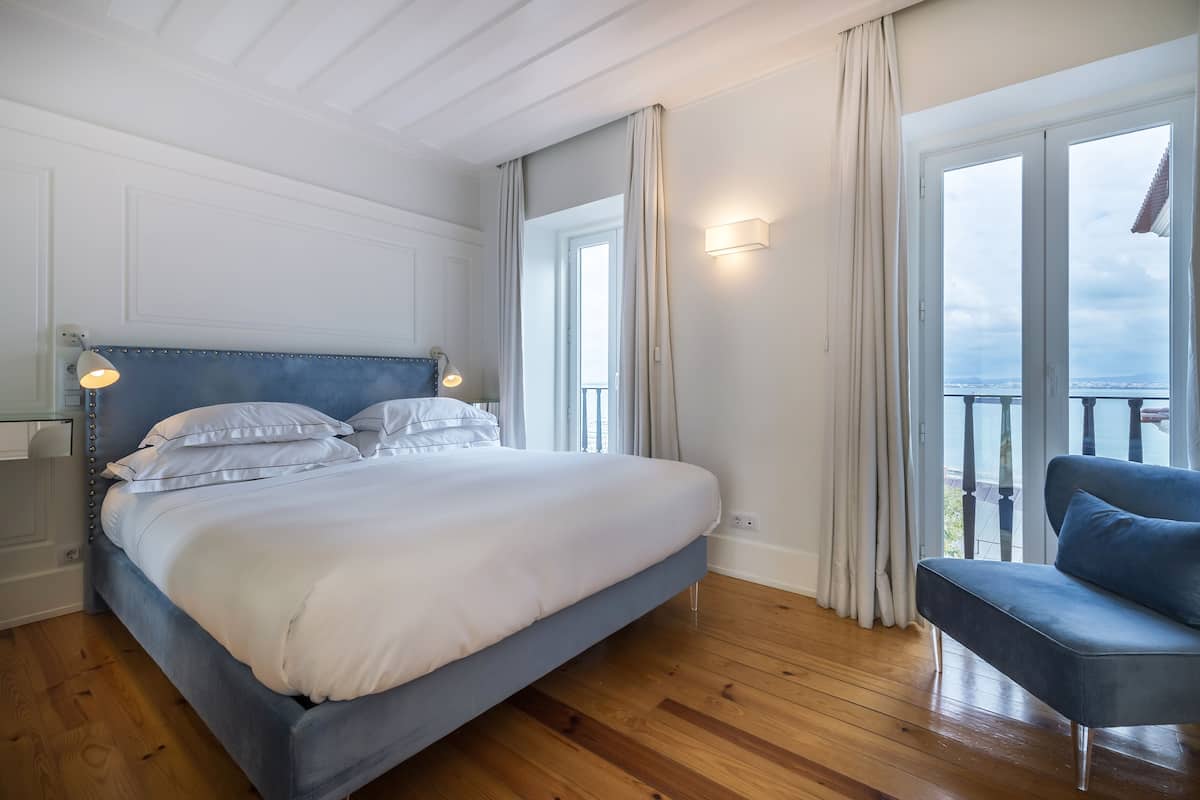 Ein Schlafzimmer mit einem großen Bett, einer blauen Kopfteil, einem Nachttisch mit Lampe, einem blauen Sessel und einem Balkon mit Blick.