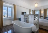 A modern bathroom with a freestanding bathtub, a large window with curtains, and a bed with a view of a building.