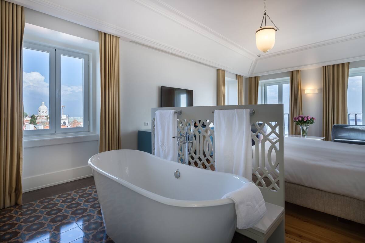 Ein modernes Badezimmer mit freistehender Badewanne, einem großen Fenster mit Vorhängen und einem Bett mit Blick auf ein Gebäude.