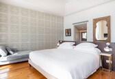 A bedroom with a large bed, a grey sofa, patterned wallpaper, and a wooden floor.