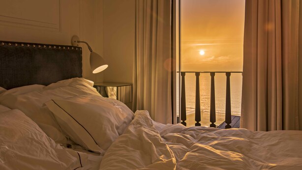 A hotel room with a bed, a nightstand, a lamp, and a view of the sunset over the ocean. - Santiago de Alfama - Boutique Hotel (Lisbon)