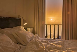 A hotel room with a bed, a nightstand, a lamp, and a view of the sunset over the ocean.